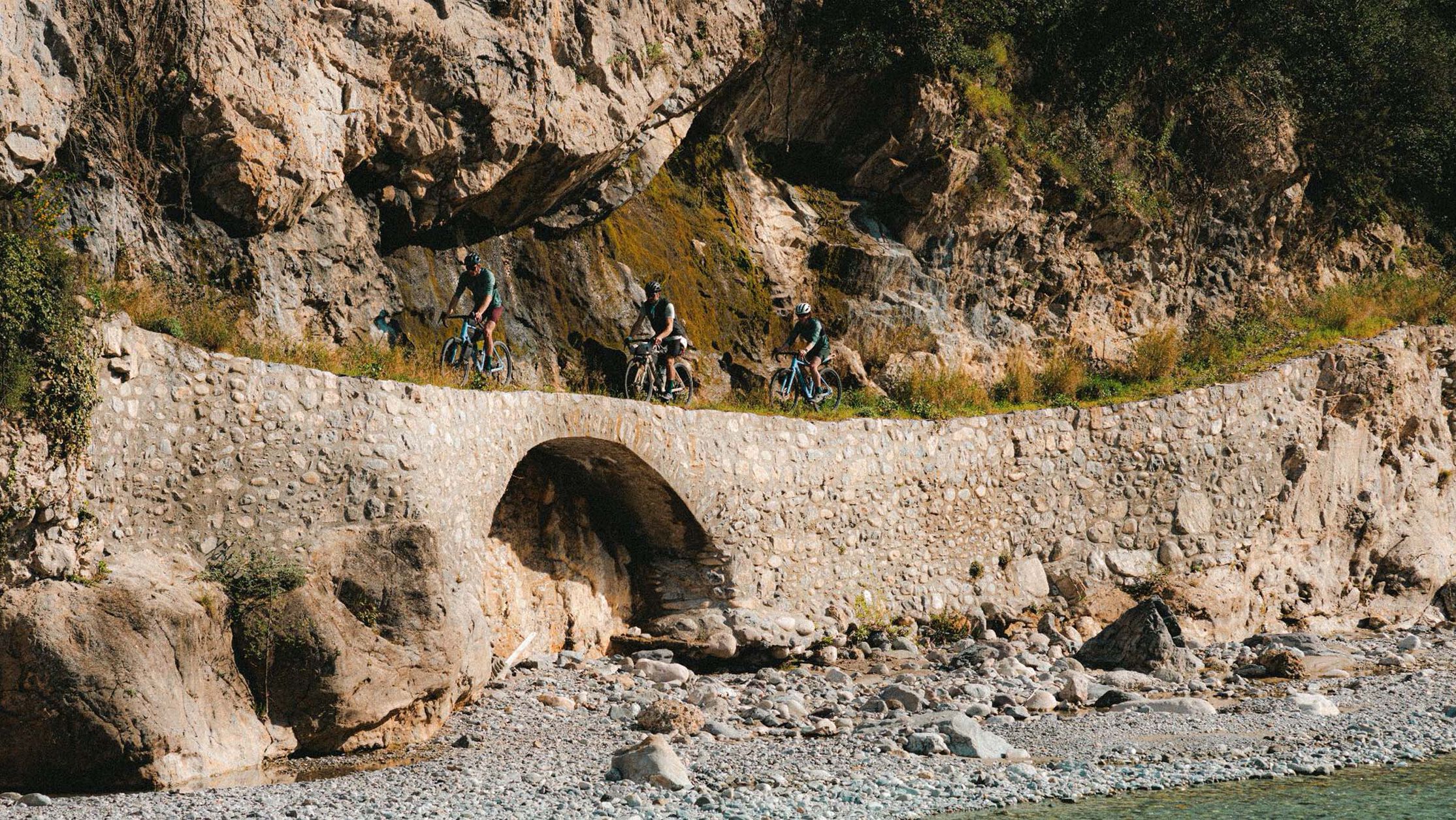 Group of cyclists riding Canyon gravel bikes across a stone bridge carved into a rocky mountainside, highlighting the bikes' off-road capability and adventure readiness.